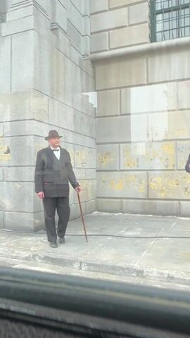 Two men dressed in suits and hats stand outside a building with a brick-like facade. The man on the left wears a black suit with a bow tie and holds a walking stick. The man on the right wears a navy suit with a tie and also holds a walking stick, gesturing with his right hand. The building has windows and a peeling yellow and gold design on the wall. The ground is paved with concrete slabs. The video appears to be shot from inside a vehicle.