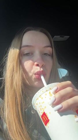 A young woman is sitting in a car drinking from a McDonald's cup. She is facing the camera and looks directly at the lens while drinking. Her lips are puckered while drinking from a white straw. She has long, straight, light-brown hair. Her nails are long and white. The car's interior is dark. Music plays in the background.