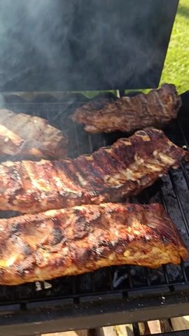 This media file captures a close-up, overhead view of ribs cooking on a grill. The ribs are positioned on a black metal grate, with visible smoke rising around them. The meat appears well-seasoned and is developing a char from the grilling process.

In the foreground, a bunch of fresh green herbs, possibly dill or parsley, is being added to the grill. A hand is visible, holding the herbs and bringing them closer to the ribs. The herbs appear vibrant and add a splash of color against the darker tones of the meat and grill.

The background suggests an outdoor setting. To the left, there is a glimpse of green grass, indicative of a yard or park. To the right, wooden elements are visible, possibly part of a fence or outdoor furniture. The lighting suggests it is daytime, likely sunny, given the brightness and clear shadows. The overall scene evokes a sense of a casual outdoor barbecue or cooking event. No visible text is present in the media file. The setting is identified as Ozyaritska-Slabadski rural council, Belarus.