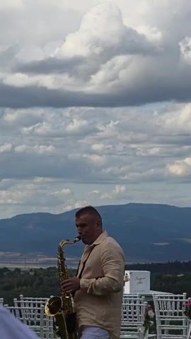 A man plays a saxophone outdoors. He is wearing a light colored shirt and white pants. He has short dark hair and is looking to the side. In the background, there are mountains and a cloudy sky. There are also rows of white chairs set up, some decorated with flowers, suggesting an outdoor event like a wedding. A white umbrella is visible in the foreground.
