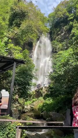 The video shows a tall, cascading waterfall surrounded by lush green trees and rocks. Several people are visible in the foreground and on a walkway leading up to the waterfall. One woman in the foreground is wearing a white patterned dress and a yellow backpack, and is looking towards the waterfall. Another person, facing away from the camera, is wearing a white t-shirt and a cap. A third person, also facing away from the camera, is wearing a denim jacket and holding a water bottle. The scene appears to be a natural attraction, possibly a park or tourist destination.