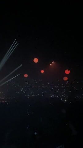 A nighttime concert is captured from a high vantage point, showcasing a vast audience in a stadium. Bright spotlights cut through the darkness, illuminating the stage. Several large, red balloons float above the crowd, contributing to the overall ambiance of excitement and spectacle.