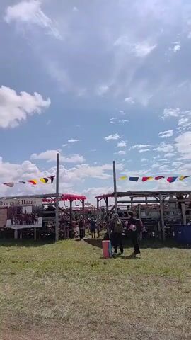 The video shows an outdoor event with temporary structures and people. There are wooden bleachers or seating areas, some covered by awnings with red trim. Flags in various colors, including red, yellow, blue, and white, are strung between poles above the structures. One sign appears to have Russian text. Several people are visible walking around and gathered in small groups. The ground is covered in grass and dirt. The sky is blue with scattered white clouds. The overall impression is of a fair, festival, or historical reenactment.