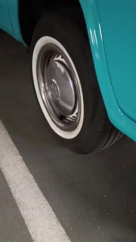 This media file showcases a vintage turquoise and white Volkswagen bus, identified by the name "Lisbeth" written in white script on its side. The bus is parked in what appears to be an indoor setting, possibly a garage or showroom, indicated by the smooth, dark asphalt floor with white painted lines and the artificial lighting.

The camera begins by focusing on the rear passenger side wheel, which features a classic chrome hubcap with a Volkswagen logo and a white-wall tire. The camera then pans forward, revealing more of the bus's exterior. We see the passenger door, the side panel, and then the front of the vehicle. The iconic round headlights, amber turn signals, and the distinctive grille of the Volkswagen bus are clearly visible. A license plate, with German plates visible, further confirms the location. The overall impression is of a well-maintained, classic vehicle. There are no people visible in the media. The lighting suggests it could be daytime or evening, but the indoor setting makes it difficult to determine the time of day or weather conditions outside. The bus is the sole focus, presented in a way that highlights its vintage aesthetic and the care taken in its upkeep.