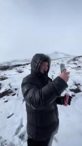 The video shows a person standing in a snowy landscape. The person is wearing a black puffy jacket with a hood and appears to be taking a picture with a phone while holding a can of Coca-Cola. The background is a snowy hillside with a mountain in the distance and a cloudy sky. The person then walks across the snowy field.