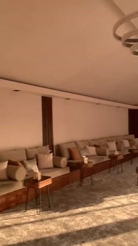 The video pans across a modern living space with a desert view outside. The room features a long, built-in sofa with multiple cushions, small side tables, and circular, multi-tiered chandeliers. The flooring is a neutral-toned, textured carpet. Natural light streams in through large windows. The camera moves to a seating area closer to the windows, revealing a view of a patio with reddish gravel, a potted flowering plant, and a hillside covered in rocks and sparse vegetation. A building can be seen on top of the hill. The focus is then drawn to the plant itself, capturing the pink flowers in detail against the desert landscape.