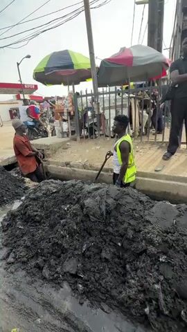 Two men are digging out a large pile of black sludge from a drainage ditch on the side of a street. One man, wearing an orange shirt, is in the ditch with a shovel. The other man, wearing a yellow safety vest over a white shirt and dark shorts, is also in the ditch with a shovel. A third man in a black shirt stands to the right, observing. In the background, there are several market stalls with umbrellas, a fence, and people walking. The ground is a mix of dirt and water. The sky is overcast.