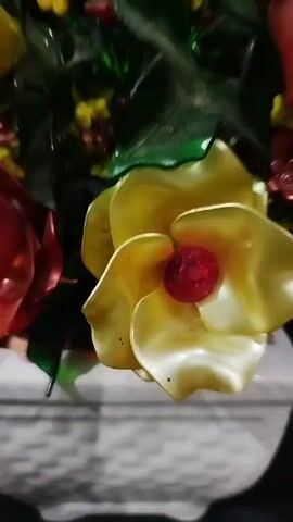 A close-up shot of a decorative floral arrangement in a white pot. The arrangement consists of artificial flowers in shades of red and yellow, with small yellow and red filler flowers. The pot is textured and rests on a black surface, likely a chair. The background is dark and out of focus. The camera slowly zooms out and pans slightly to the left, revealing more of the floral arrangement and the black chair it is placed on. The background remains dark and indistinct. As the video progresses, the focus shifts slightly to the entire arrangement.