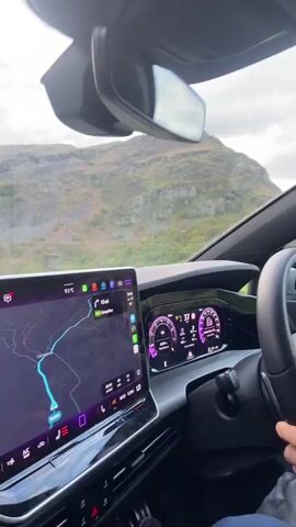 The video shows a driver's perspective of driving along a winding road. The dashboard of the car is visible, showing a navigation screen with a map. The scenery outside the window consists of rolling green hills and mountains. The sky is overcast with clouds. The video is taken during daytime.