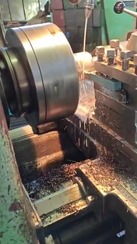 A close-up, side-view shot shows a metalworking lathe in operation. A large, metallic workpiece is held in a chuck on the left, rotating rapidly. A stream of coolant is being sprayed onto the workpiece from an articulated nozzle positioned above and to the right. A cutting tool, mounted on the lathe's tool post, is seen removing material from the workpiece, creating shavings that fall into a trough below. The trough is filled with accumulated metal shavings and coolant. The background is out of focus, appearing to be a workshop environment with a green tarp or curtain visible behind the machinery. The video focuses on the precision machining process.