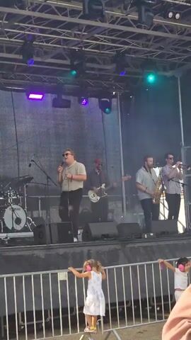 A live band performs on an outdoor stage under a bright sky. A lead singer in a light-colored collared shirt and dark pants stands center stage, holding a microphone and singing. To his left, a drummer is seated behind a drum kit. To his right, a guitarist plays a white electric guitar. Further to the right, a saxophonist plays a tenor saxophone, and behind him, a trumpeter plays a trumpet. The stage is equipped with lighting rigs overhead, featuring purple and green lights. In the foreground, a metal barricade separates the stage from the audience. Two young girls stand near the barricade, one in a white dress and the other in a pink top, both appearing to be watching the performance. The overall environment suggests a daytime festival or outdoor concert.