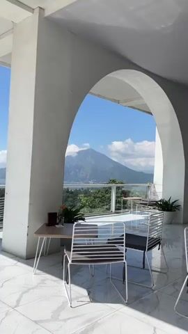 The video shows an outdoor seating area with a view of a volcano and a lake. There are several tables and chairs set up on a tiled patio. In the foreground, there are two tables with chairs, each with a drink on it. In the background, a large volcano is visible, with a lake and mountains beyond it. The sky is blue with some white clouds. The sun is shining, indicating it is daytime. The overall impression is a scenic and peaceful location, likely a cafe or restaurant.