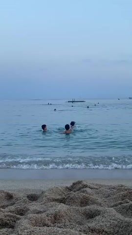 This is a video of a beach scene at dusk. The camera is positioned on the sand, looking out towards the ocean. In the foreground, there is a patch of textured sand with small waves lapping at the shore.

The water is calm and a clear turquoise color near the shore, becoming a deeper blue further out. Several people are in the water, swimming and enjoying the evening. In the middle ground, two children are visible, one facing the camera, the other with their back to it. Further out in the water, other individuals are scattered, some appearing to be swimming or floating.

In the background, there is a wooden pier or platform extending into the water, with a few people on it. A white boat with a red stripe on its hull is sailing in the distance to the right, and another smaller white boat is visible near the pier. The sky is a soft gradient of pale blue and lavender, indicating it is either sunrise or sunset. There is no visible text in the video. The overall mood is peaceful and relaxed, suggesting a pleasant end to a day at the beach in Arenys de Mar, Spain.