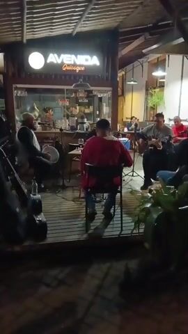 The video shows a live band performing at night in what appears to be an outdoor area of "Avenida Quiosque." A man wearing a red jacket is sitting in a chair facing the band. To the left, a percussionist is playing a tambourine, and a guitar case is leaning against the wooden structure. A guitar player is seen on the right along with some other people. The area is lit by warm overhead lights and there is a small snack stand behind the musicians. Several patrons are eating at tables in the background. The environment seems relaxed and lively with the band providing musical entertainment.