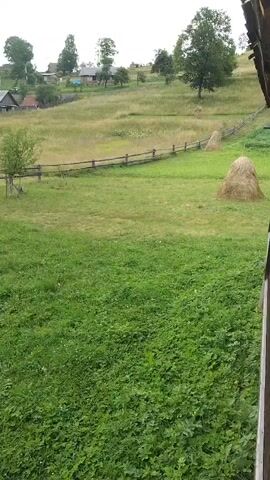 The video shows a rural landscape with rolling hills covered in green grass. In the distance, there are several houses with dark roofs and a wooden fence runs along a path. There are also haystacks in the field. The sky is overcast and the overall impression is one of a peaceful, rural setting. The video is static and there is no movement.