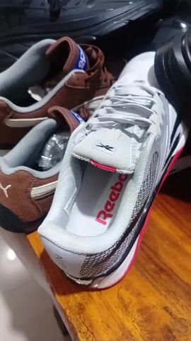 The video displays a collection of shoes on a wooden table, likely in a store or a dedicated display area. In the foreground, a pair of white and silver Reebok sneakers with bright pink accents on the sole are prominently featured. The word "Reebok" is visible in pink text on the insole of this shoe. Next to them, a pair of brown suede Puma sneakers, with the Puma logo on the side, are also visible. Several other pairs of shoes, predominantly black sneakers and dress shoes, are scattered on the table. In the background, shelves are stacked with numerous other sneakers of various colors and designs, and a brown checkered bag is also seen. A hand briefly enters the frame at the bottom right. The setting suggests an indoor environment, possibly a retail outlet given the quantity and display of footwear.