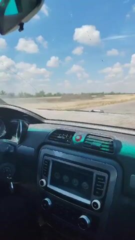 The video provides an interior view of a car, focusing on the dashboard and the windshield. The car appears to be in motion on a track or a road, with a landscape of dry vegetation and a bright blue sky with scattered clouds visible through the windshield. The dashboard is dark and features a central infotainment system with a screen displaying four circular icons. To the left of the screen are two knobs, and to the right are several buttons. Above the screen are air vents, one of which has a red and blue accent. The car's rearview mirror is visible at the top of the frame. The camera perspective suggests the video is taken from the passenger seat or a fixed position within the car. The car seems to be navigating a turn, as indicated by the slight tilt of the vehicle and the blur of the passing scenery. There is a visible crack on the windshield. Dust or debris is kicked up along the side of the road.