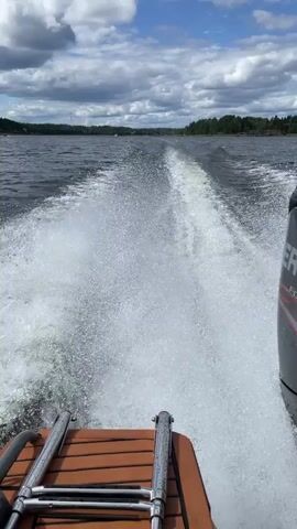 This is a first-person perspective video of a boat moving through water. The camera is positioned at the back of the boat, looking forward. The boat is creating a large wake behind it. The water is dark and appears choppy. On the left side of the frame, the edge of the boat is visible, with a wooden surface and a metal ladder. On the right side, a black boat engine is prominently displayed with the word "MERCURY" and "FOURSTROKE" printed on it. The background shows a tree-lined shore under a cloudy sky with patches of blue. The overall impression is of a fast-moving boat on a lake or a large body of water.