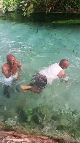 Two men are swimming in a clear, turquoise-colored body of water. The water appears shallow, with visible rocks and vegetation at the bottom. The man closest to the camera is mostly submerged. The other man, wearing a white tank top, is swimming and then splashes into the water, submerging himself completely. The surrounding environment includes green plants and trees, indicating a natural setting. There is exposed wood on the left bank.