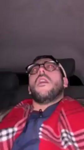 A man with a beard and glasses is sitting in the back seat of a car, wrapped in a red and white plaid blanket. He is looking up and singing along to a song playing. The car's interior is visible and it is dark outside.