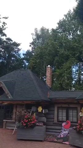 The video pans across the exterior of a rustic wooden building with a dark green roof. The walls are constructed from large logs. The building features several windows and an open doorway. There are decorative elements such as yellow flowers attached to the logs and cutouts of cartoon characters, including a mouse wearing a red hat and two rabbits. A brick chimney is visible on one side of the building. The ground in front of the building is paved with red brick. Tall trees with green foliage are visible in the background. The sky appears overcast.