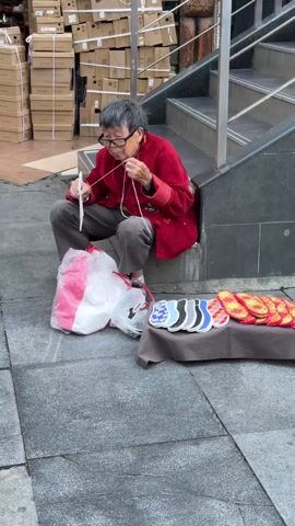 An elderly Asian woman in a red jacket and glasses sits on concrete steps outside and sews a black and white shoe. A plastic bag is to her left. In front of her is a small table on which several colorful shoe inserts are laid out. Behind her is a stack of tan boxes and a set of metal stairs. The pavement is gray.