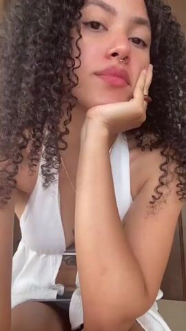 A medium close-up shot shows a woman with curly dark hair and a nose ring. She is wearing a white halter top with a low neckline, revealing a tattoo on her chest. She rests her chin on her hand and makes a puckered expression. The background is blurred, but appears to be an indoor setting.