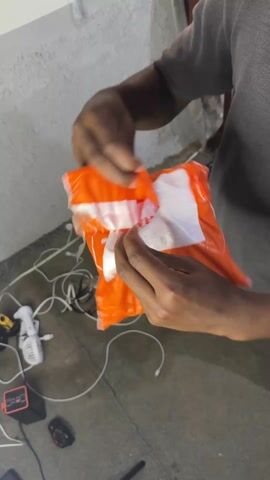 A person wearing a grey t-shirt and blue shorts is opening an orange and white delivery package. The package is opened to reveal an item wrapped in clear plastic. The person removes the item from the package. The floor is grey and cluttered with cables, chargers, a camera, and other miscellaneous objects.  The wall behind the person is white and textured.