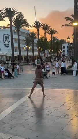 A man dressed in a loincloth and what appears to be a tribal headdress is performing in a pedestrian area. He is holding a long stick and appears to be in motion, possibly dancing or performing a trick. In the background, there are palm trees, buildings, and a crowd of people walking. The sky is a warm orange and yellow, suggesting it is either sunrise or sunset. The ground is paved with stone tiles. The man is barefoot.