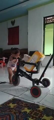 A toddler is standing next to a tricycle with a package on its seat. The tricycle has a parent handle. The toddler is wearing a white tank top and a diaper. The tricycle is black and red and has three wheels. The package is yellow and white and appears to be diapers. The toddler is leaning forward and touching the tricycle. The scene is indoors in a room with tiled floors. There is furniture in the background, including sofas and chairs. There is also a window and a door. The wall has a red curtain and a picture of a bird. The ceiling is blue.