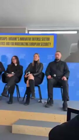 A video shows a panel discussion with five speakers. Four speakers are sitting on a small stage, and one speaker is on a video screen visible in the background. There are two men on the left and three women in the center of the group of speakers on stage. The man on the right of the stage is wearing all black and has a microphone in his hand. The woman to his left is wearing a black dress and boots. The other two speakers are dressed in black suits. The speaker on the far left of the stage is wearing a grey suit and holding a piece of paper. A banner is visible in the background that reads, “TECH DEMOCRACY PARTNERSHIPS: UKRAINE’S INNOVATIVE DEFENSE SECTOR IS VITAL FOR NATIONAL DEFENSE AND FOR MODERNIZING EUROPEAN SECURITY.” In the foreground, a person is visible in the lower right corner of the video. There is a chair with the word “RESERVED” printed on it visible in the bottom left corner of the video.