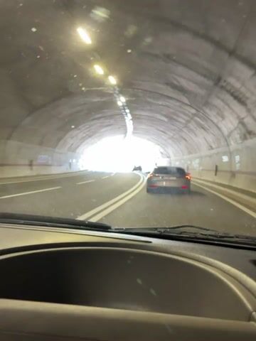 The video begins inside a car, looking forward through the windshield as it drives through a tunnel. The tunnel is illuminated by overhead lights, and the end of the tunnel, showing a bright, sunny sky and green hills, is visible in the distance. A silver car is ahead, also heading towards the exit. As the car exits the tunnel, the scene transitions to a sunny day on a winding road. The car continues to follow the silver car, which is now on a highway with a steep, rocky hillside covered in green vegetation to the left and a guardrail to the right. The sky is a clear, bright blue. Several streetlights are visible along the side of the road. Other vehicles, including a truck, are also on the road in the distance. The video appears to be captured in the daytime under clear weather conditions. The license plate of the silver car is visible and reads "PG NP 246".