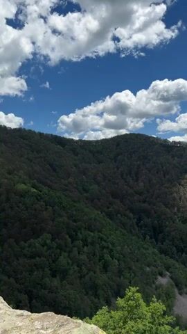 The video pans across a verdant mountainside covered in trees under a bright blue sky with fluffy white clouds. The slope is densely forested with lush green foliage. In the distance, more rolling hills and mountains are visible, some with patches of lighter green or brown, suggesting variations in vegetation or terrain. The foreground shows the edge of a rocky outcrop, providing a viewpoint overlooking the expansive landscape. The overall impression is a natural, outdoor scene, likely a scenic vista from a hiking or viewing point. There are no people or identifiable objects other than the natural environment.