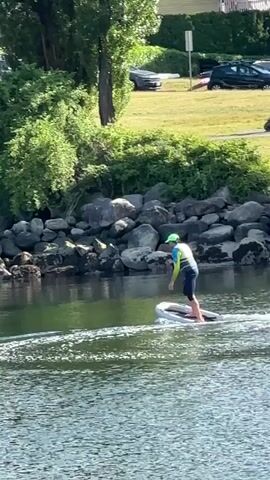 A person wearing a blue swimsuit, yellow and green life vest, and a green hat is on an electric surfboard in a body of water. They are crouched on their knees, facing forward, and appear to be moving. The water is calm with some ripples behind the surfboard. The background shows a rocky shoreline with green trees and bushes. Further in the background, there are cars and people visible on a grassy area. The lighting suggests it is daytime.