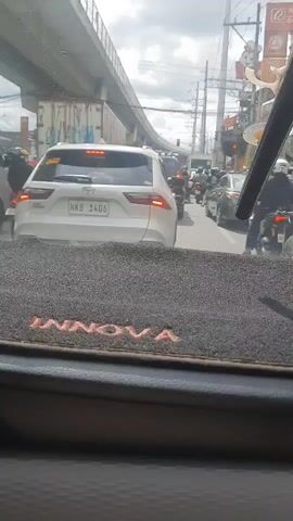 The video is a point of view shot from inside a vehicle looking forward at a congested city street. In the foreground, the dashboard of the vehicle is visible with a dark gray mat that has the word "INNOVA" embroidered in orange. Beyond the dashboard, a white SUV with license plate NKB 1406 is in the lane ahead. A traffic light is visible further down the road, as well as numerous other vehicles including motorcycles and cars. Overhead, a large elevated highway structure is visible, supported by concrete pillars. Power lines and street signs are also present, including a sign that appears to be for a fast food restaurant. The sky is overcast. There are no discernible actions or interactions occurring within the video.
