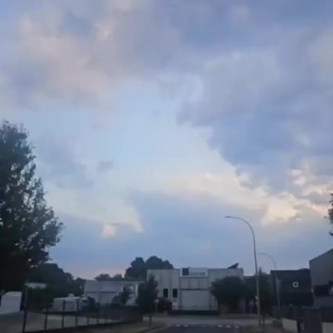 The video shows a street scene with several buildings and trees. The sky is cloudy. There are streetlights and a traffic sign. The buildings appear to be industrial or commercial. In one shot, there are three flags with logos on them. In another shot, there is a sign that says "alosa." The video is shot from a moving vehicle, as indicated by the slight blurring of the scene.