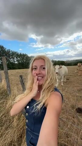The video shows a woman with long blonde hair and a blue denim outfit holding her hand to her mouth and smiling. She is in a field of dry grass with a fence in the foreground. Behind her, a white cow stands looking at the camera, with several other cows lying down in the field further in the background. The sky is cloudy and gray. The camera pans across the field, showing more cows and a line of trees in the distance.