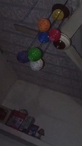 The video shows a ceiling fan with a decorative light fixture hanging from it. The light fixture is comprised of several colorful, spherical lamps arranged in a cluster. The lamps are various colors including yellow, orange, blue, red, purple, green, and white. The ceiling has a textured, patterned design. Below, on a shelf, there are various boxes, a small lamp, and other items. The overall lighting in the room is dim.