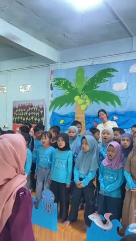A group of young children, most wearing matching light blue shirts and some wearing hijabs, stand on a raised platform in front of a blue banner decorated with a palm tree, the sun, and clouds. The banner also has text that appears to read "PERPISAHAN IES SCHOOL ANGKATAN KE 8". The children appear to be singing or performing, with some holding microphones or gesturing. A woman in a white top stands behind the children, looking towards them. The environment appears to be an indoor room with a ceiling light fixture visible. In the foreground, there are some cardboard cutouts of sea creatures, including an octopus and a dolphin. The overall atmosphere suggests a school event, possibly a graduation or farewell ceremony.