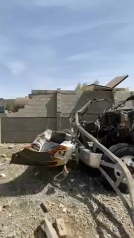 The video shows the aftermath of an explosion and fire in what appears to be an industrial or residential area. The first shot reveals the charred remains of a tanker truck, with its front end heavily damaged and a portion of the cabin burned away. Behind it stands a damaged brick wall. The camera pans to show the full extent of the tanker truck, which is burnt and has a bullet hole in the tank. A man with a beard stands near the wreckage. The final shot shows the site of a large explosion and fire. A plume of black smoke billows into the sky, flames are visible, and the surrounding area is in ruins.
