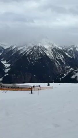 The video shows a snowy mountain landscape under a cloudy sky. In the foreground, there's a snow-covered slope with ski tracks. A safety fence is visible, along with some small structures. Two people are skiing on the slope. The background features a large, snow-covered mountain peak, partially obscured by clouds.