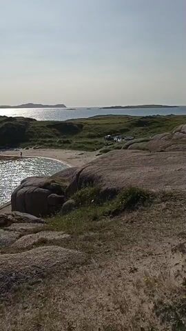 The video shows a coastal scene with several islands in the background. The foreground consists of rocky terrain with patches of grass. In the middle ground, there is a small sandy cove with clear turquoise water. Several people are visible on the beach and in the water, some swimming and others standing. A small white boat is docked on the left side of the cove. Further out in the water, there are other boats, including a larger white boat with people on board and several smaller boats with individuals paddling in them. On the shore in the distance, there is a pier with several cars and people gathered. The water is calm and reflective, with sunlight shimmering on its surface. The sky is clear and blue, indicating a sunny day. The overall impression is a serene and idyllic coastal location, likely a popular spot for recreation.