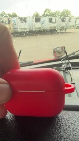 A hand is holding a red charging case for wireless earbuds. The case is rounded and has a small loop on the side. The background shows a blurry view through a window, with rain on the glass. Several white refrigerated trucks are visible in the distance, parked in a lot. A pair of glasses is resting on the dashboard in front of the case. The overall environment appears to be inside a vehicle.