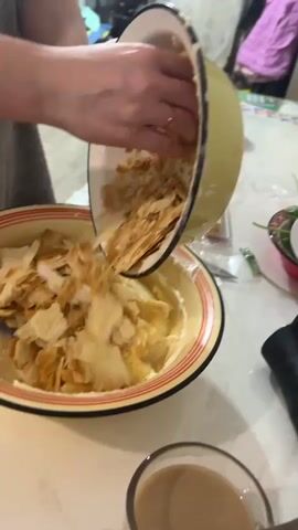 The video shows a person in a close-up shot, preparing a dessert. The person is holding a bowl in their left hand and a spatula in their right hand, using the spatula to scoop pieces of layered pastry from the bowl into a larger, yellow-rimmed bowl filled with a creamy white substance. The pastry pieces are golden brown and appear to be broken into irregular shapes. The larger bowl already contains a significant amount of pastry pieces mixed with the cream. In the background, out of focus, is a room with some clothing hanging on a rack and other kitchen items on a table. The lighting suggests an indoor setting, possibly a kitchen. The action is focused on the preparation of the dessert, with the person actively mixing the ingredients. There's a glimpse of a glass with a beverage in the foreground at the bottom right corner of the frame.