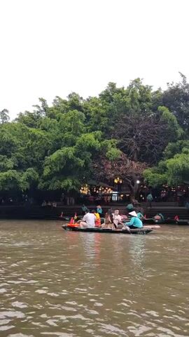 The scene captures a lake with several boats carrying people, set against a backdrop of lush green trees under a cloudy sky. Most of the boats appear to be small, round Vietnamese boats. People are wearing casual attire, and some are wearing conical hats, a traditional Vietnamese headwear. Some people are wearing life vests. The boats are moving on the water, with some individuals rowing, while others seem to be enjoying the ride. A structure that looks like a shop or restaurant is faintly visible on the shore amidst the trees. The water surface is slightly choppy, reflecting the ambient light and suggesting a gentle breeze.
