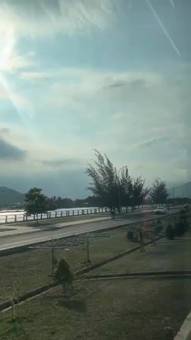 The video shows a coastal road with trees and grass. The sun is shining brightly, and there are some clouds in the sky. Several cars and a truck are driving on the road. The video appears to have been filmed from a moving vehicle. The overall scene is one of a sunny day in a tropical or subtropical location, possibly Vietnam as indicated by the text overlay.