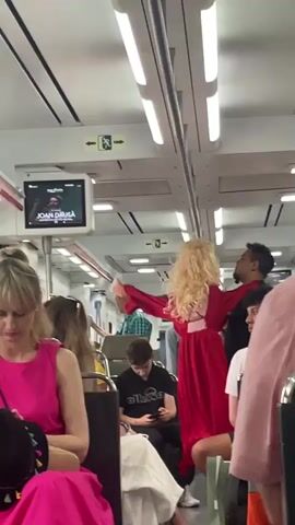 The video captures a dynamic scene inside a modern train or metro car, likely in Sant Andreu de Llavaneres, Spain. In the center of the aisle, a woman with long, curly blonde hair, wearing a flowing red dress, is performing or interacting intimately with a man in a black shirt and pants. The man holds the woman close, leaning in towards her as if singing or engaging in a romantic dance, while she gestures with her arms, her back mostly to the camera.

Numerous other passengers occupy the train car, some seated and some standing. In the foreground on the left, a woman in a bright pink top is visible, looking downwards. A young man wearing a black t-shirt with an "ellesse" logo is seated in the middle, focused on his phone. A man with a bald head and a checkered shirt stands further back. Other individuals are scattered throughout, with varying levels of attention paid to the central performance.

Above the passengers, a screen displays changing names of musical artists, including "JOAN DABSA," "ANTONIO JOSÉ," "MIGUEL RÍOS," and "LEIRE MARTÍNEZ + MARLENA," suggesting the playback of music videos or advertisements. The train interior is brightly lit by overhead fluorescent lights, with white walls, overhead luggage racks, and green emergency exit signs visible. The atmosphere is one of lively, impromptu entertainment within a public transportation setting.