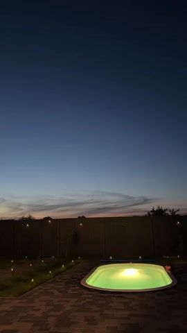 The video displays an outdoor scene at dusk or night, with a bright green illuminated swimming pool as the central focus. The pool is set in a paved area and surrounded by a lawn with small, strategically placed lights. A wooden fence forms the background, with scattered trees and the twilight sky visible above. The sky transitions from a deep blue overhead to lighter shades near the horizon, where wispy clouds are present. No people or animals are visible in the frame.