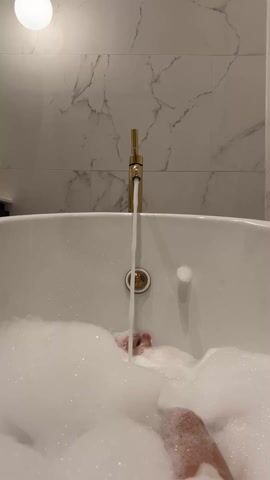 A person is relaxing in a bathtub filled with a lot of fluffy white bubbles. A gold-colored faucet is running, with a stream of water flowing into the tub. Only parts of the person's legs and feet are visible through the bubbles. The bathtub is white and the background features white marble-style tiles. A round light fixture is attached to the wall above the tub.