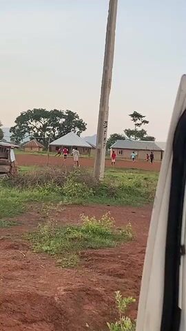 The media file captures an outdoor scene in Firse, Nigeria. In the foreground, the edge of a white vehicle door is visible on the right, suggesting the footage is from inside a vehicle looking out. The ground is a reddish-brown earth, with patches of green grass and some dry brush. A tall, grey utility pole stands prominently in the middle of the frame, casting a shadow.

Beyond the immediate foreground, a group of people are engaged in what appears to be a game of football (soccer) on a dusty field. Several individuals, mostly young men and boys, are dressed in casual clothing, with some wearing red shirts. They are spread across the field, running and interacting with a ball that is not clearly visible. In the background, there are several simple structures, likely houses, with thatched or corrugated metal roofs, typical of a rural Nigerian setting. A large tree with a wide canopy is situated to the left of the frame, and other trees and vegetation are scattered throughout the landscape. Distant hills or mountains are faintly visible on the horizon.

The time of day appears to be late afternoon or early evening, given the soft, diffused light and the long shadows cast by the utility pole and the structures. The sky is a pale, clear blue, suggesting fair weather. The overall atmosphere seems relaxed and communal, with the focus on the playful activity of the people. There is some faint, possibly handwritten text on the utility pole, but it is too indistinct to be read. The scene depicts a common everyday activity in a rural Nigerian community.
